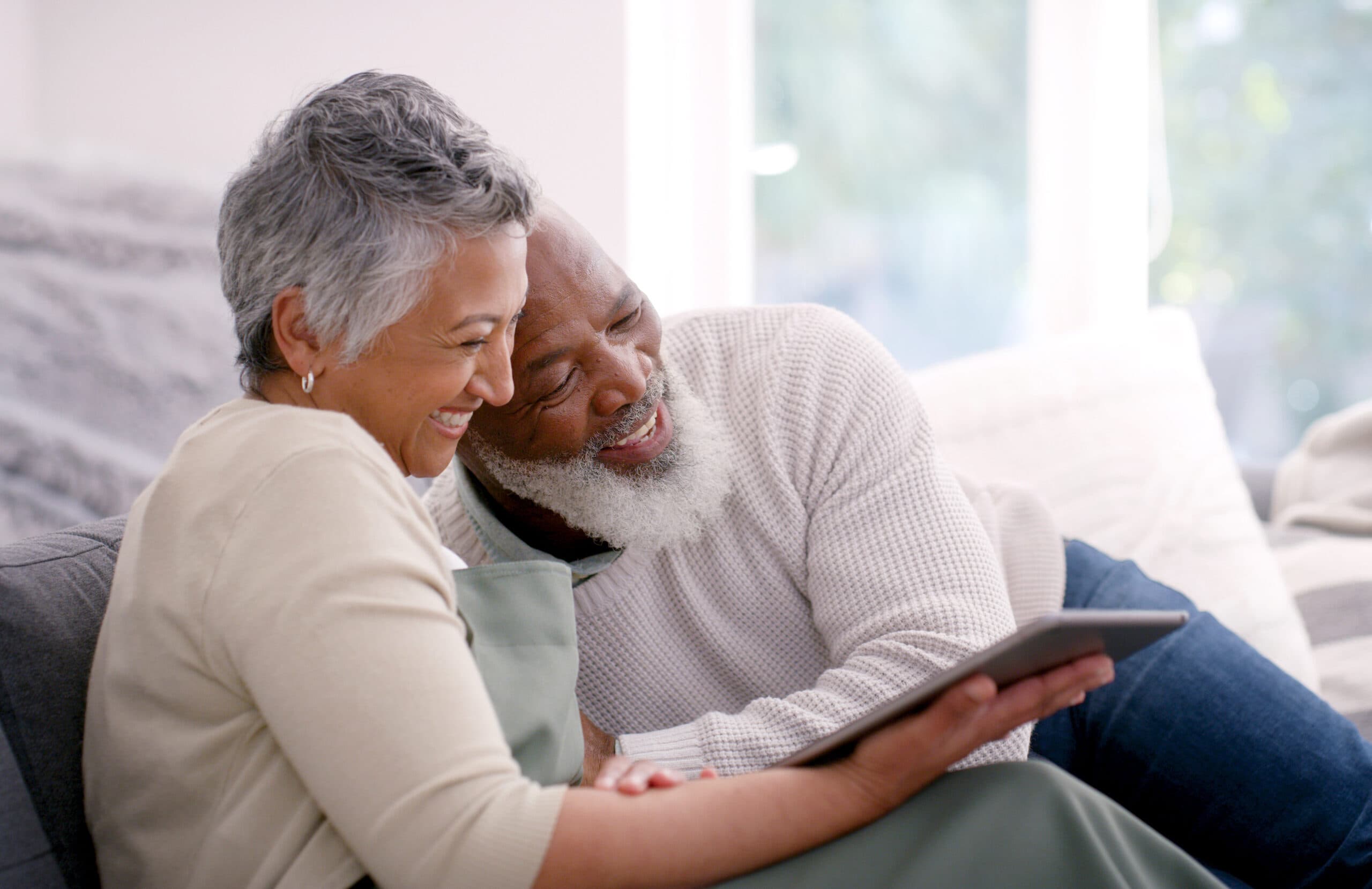 A happy couple watches a show on a tablet, cozy on the couch.