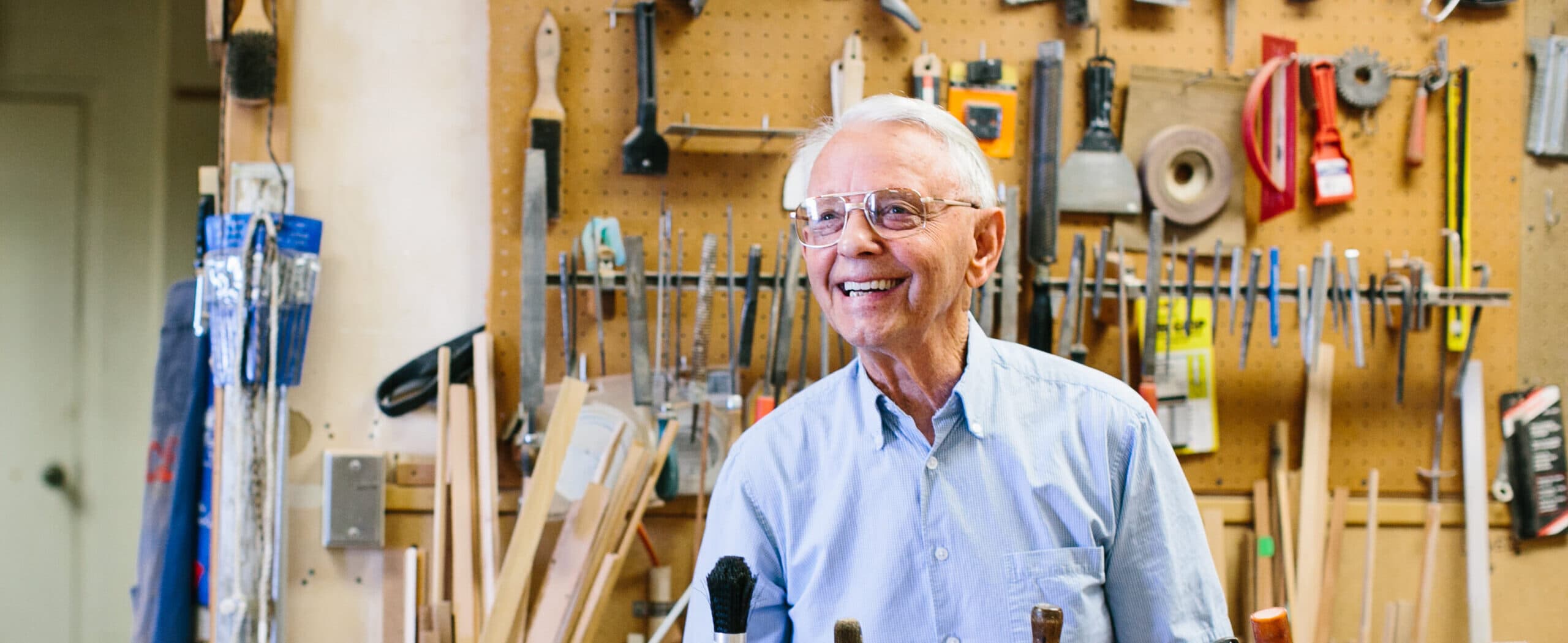 A smiling man works in a woodshop, surrounded by tools.